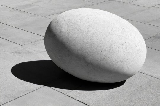 A large, smooth, egg-shaped stone sculpture rests on a paved surface, casting a deep shadow