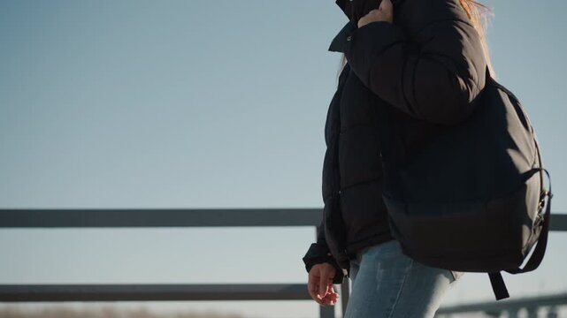Woman wearing black puffer jacket and carrying large dark backpack walks outdoors under clear blue sky with metal railing in background. Her long hair is visible as woman moves maintaining