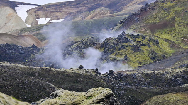 Steaming geothermal hot springs with colorful moss-covered mountains and lingering snow patches rise in the Icelandic highlands, with hikers exploring the geothermal area.