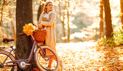 Beautiful woman sits in clearing surrounded by fallen leaves, reading book at sunset. Woman enjoys reading while sitting by tree with bicycle in autumn forest. Concept of leisure and enjoyment. © maxbelchenko