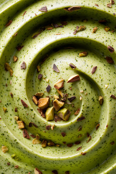 A top view close up of smooth swirling green pistachio paste texture sprinkled with chopped crunchy pistachios and nuts bits creating an organic dessert background