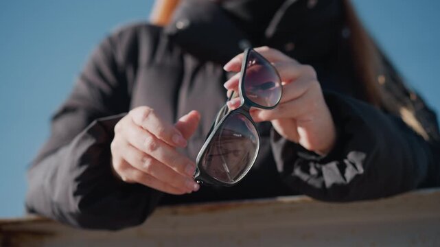 Woman holds and examines pair of sunglasses adjusting them in her hands against outdoor backdrop. Her focused interaction with eyewear is captured as woman inspects details like frame shape
