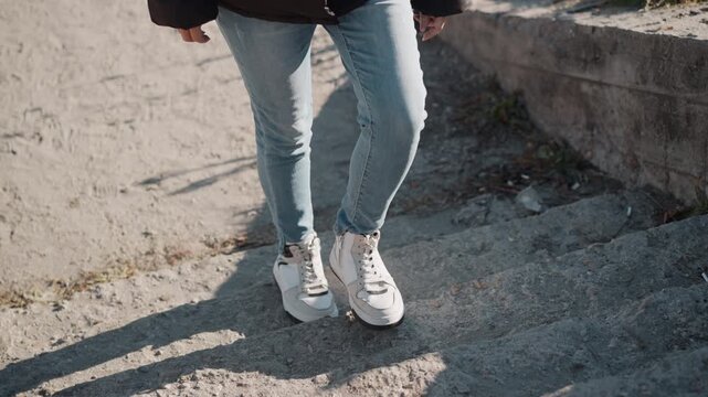 Woman walks up concrete steps outdoors wearing light blue jeans and white sneakers. focus remains on her lower body as woman ascends stairs in natural daylight with textured background