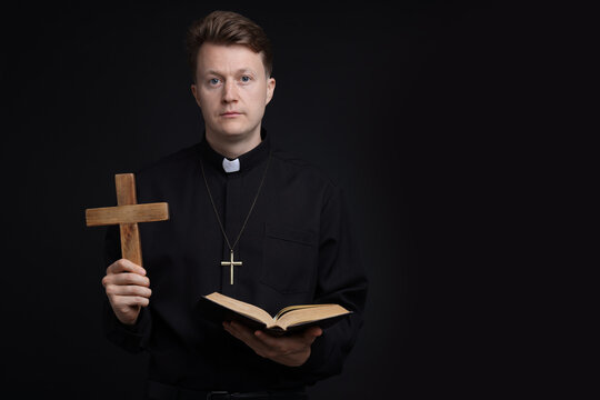 Priest in cassock with wooden cross and Holy Bible on black background, space for text