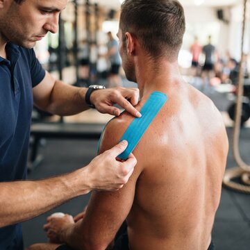 Physical therapist applying blue kinesiology tape to shoulder of male athlete in gym.