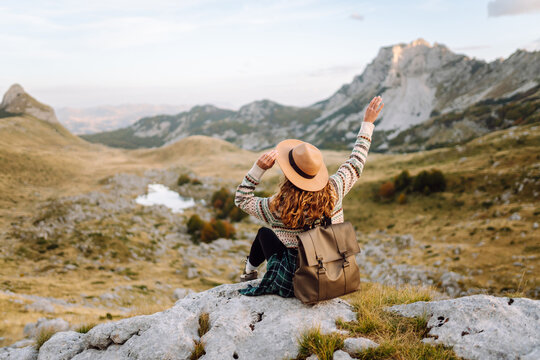 Smiling traveler wearing hat and backpack sits on rocks in mountains at sunset. Woman is enjoying mountain landscape while hiking outdoors. Concept of beauty, nature, and travel.