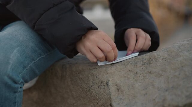 Woman wearing black jacket and blue jeans sits on stone handling white paper. Her hands manipulate folded material against outdoor background with soft focus. scene emphasizes calm