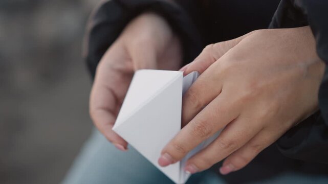 Woman holds and examines white paper origami boat gently rotating it in her hands to display its folded structure. Her fingers with neatly manicured nails interact carefully around small