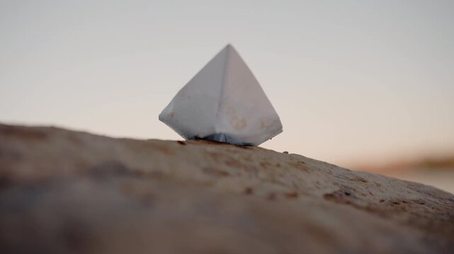 White paper pyramid rests steadily on textured rock surface under soft warm lighting. scene maintains consistent composition with geometric shape as focal point against natural background