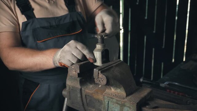 A master technician in gray overalls operates a hand tool on a metal workpiece secured in a vise inside a workshop with wooden walls and various tools in the background