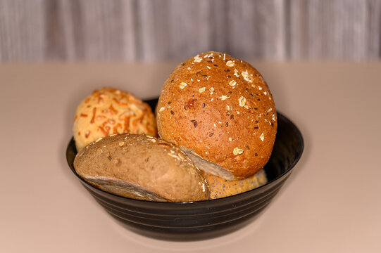 A bowl holds several types of bread rolls. The rolls show different colors and textures. They sit on a smooth, neutral-colored surface in a bright setting