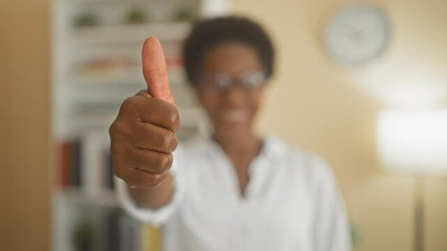 Woman showing thumbs up with prominent thumb extended toward camera in a building interior with bookshelf and clock visible; warm approval.