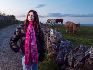 Young woman stands beside stone wall, wearing pink scarf and black jacket. She gazes calmly into distance, hands clasped in front. Cow grazes peacefully nearby on grassy hillside © mark_gusev