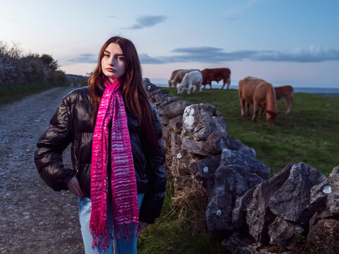 Young woman stands beside stone wall, wearing pink scarf and black jacket. She gazes calmly into distance, hands clasped in front. Cow grazes peacefully nearby on grassy hillside