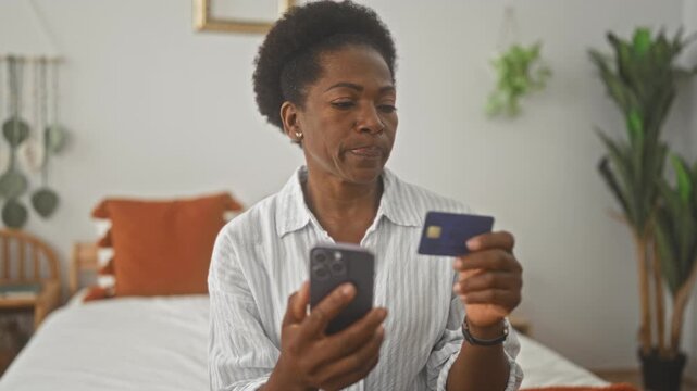 Woman holding smartphone and creditcard on a bed, checking card details and tapping screen in a building; concern financial planning.