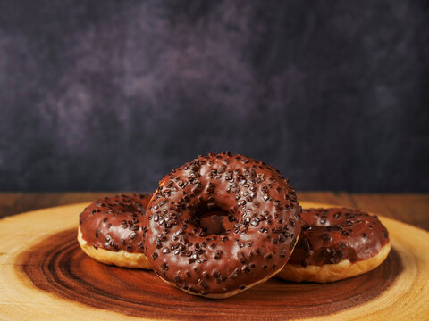 Three chocolate-glazed donuts sit on a wooden plate. Purple light reflects off sugary sprinkles and glossy glaze. Warm tones contrast with cool background hues. Perfect for food blogs, dessert ads
