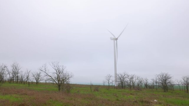 Wind turbine rotating on a foggy rural landscape. Single wind turbine generating clean, renewable energy with its blades rotating on a green hill during a foggy and overcast day