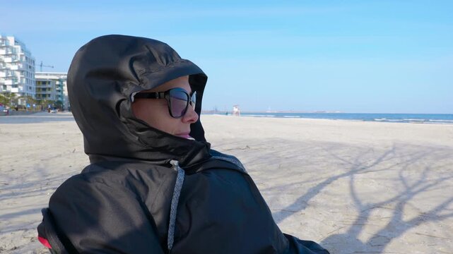 Woman relaxing on a sandy beach. Pensive woman in a black hooded jacket and sunglasses sitting on a sandy beach, looking out at the calm sea on a sunny but cool day, enjoying a moment of solitude