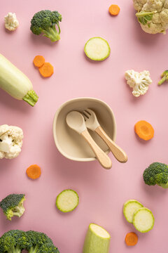 Baby first feeding with bowl and wooden spoon surrounded by fresh vegetables on vertical pastel pink background