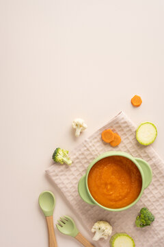 Baby's first feeding: healthy carrot puree in a green bowl with wooden spoon and fresh vegetables on vertical beige backdrop