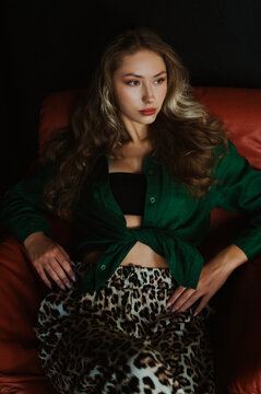 Fashionable confident woman wearing stylish summer outfit with green linen shirt, crop top, leopard print trousers, posing, sitting on brown leather sofa. Fashion portrait. Natural day light