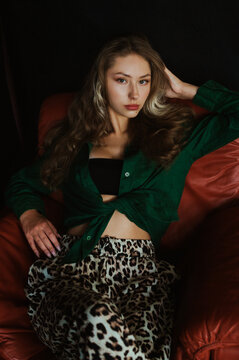 Fashionable confident woman wearing stylish summer outfit with green linen shirt, crop top, leopard print trousers, posing, sitting on brown leather sofa. Fashion portrait. Natural day light