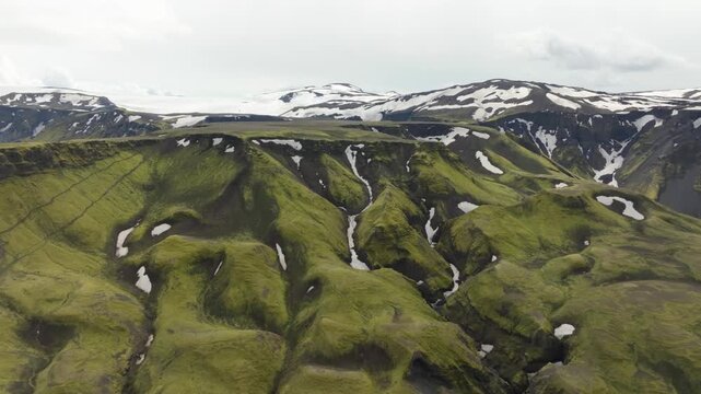 Aerial drone shot shows the Fimmv&ouml;r&eth;uh&aacute;ls hiking trail on Iceland. The drone pushes in, booms up, and pans left revealing stunning green volcanic mountains, snow patches, and a glacier.
