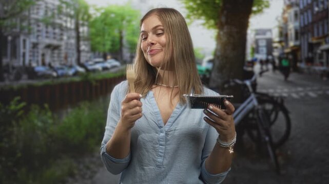 Woman holding wooden fork and meal container on sunlit street with smiling expression; culinary pleasure.