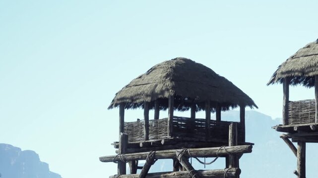 Two rustic huts made of wood and thatch rise above the shoreline, surrounded by mountains and a vast open sky. A tranquil scene reflecting simplicity and natures beauty.