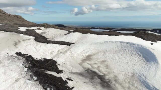 Aerial push in drone shot reveals a hiker traversing the Fimmv&ouml;r&eth;uh&aacute;ls trail on Iceland, surrounded by a vast, remote volcanic landscape with snowfields extending to the distant ocean.