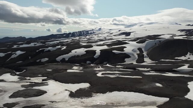 Drone pans left over the Fimmv&ouml;r&eth;uh&aacute;ls hiking trail on Iceland, revealing a vast, rugged volcanic landscape dotted with snowfields and a distant glacier under a soft light sky.