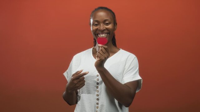 Young african american woman smiling while holding a small red heart and pointing finger to it against a warm orange studio wall; joy connection.