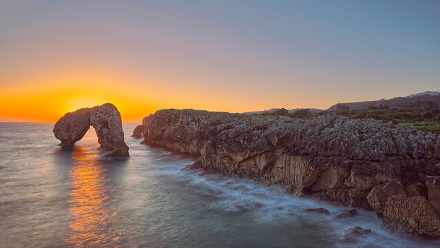 Spectacular sunrise over Castro de las Gaviotas islet, rock arch next to a limestone cliff, backlit by the orange sunlight. Llanes-Asturias-Spain-087