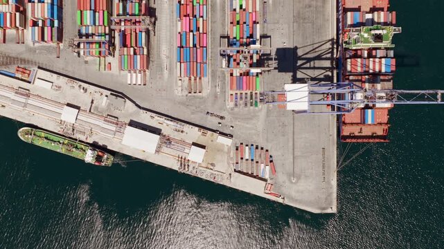 Top-down aerial view of a large gantry crane moving containers from a docked cargo ship in a busy industrial harbor, showing the global shipping and international logistics industry