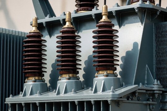 Closeup on three phase distribution transformer bushings with porcelain insulators and metal parts. Electrical component for power supply network, electric infrastructure, energy transmission.