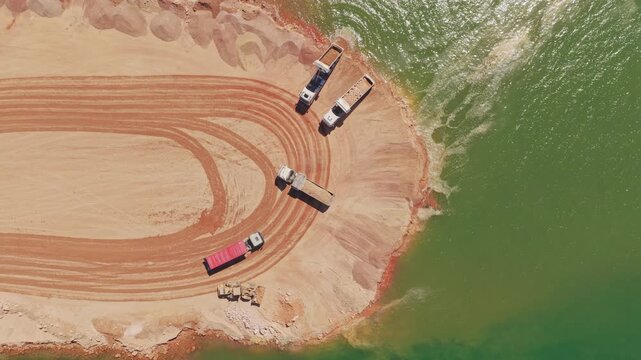 Top-down aerial view of industrial dumper trucks unloading sand into a lake at a construction site, creating a new shoreline and expanding the land area for development