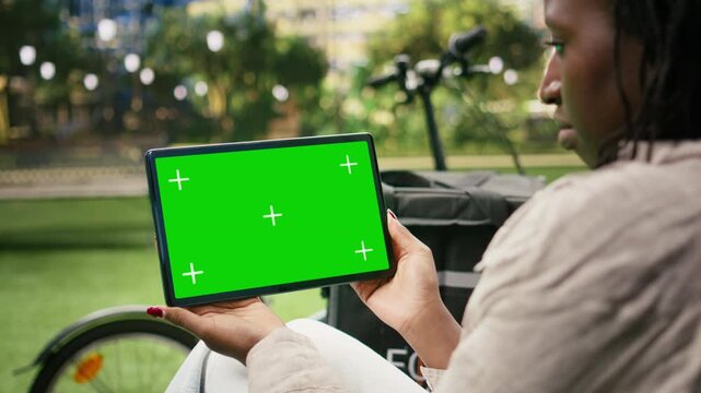 Green screen chroma key appears on a tablet held by a woman courier in a city park, providing a replaceable widescreen display for web and advertising content.