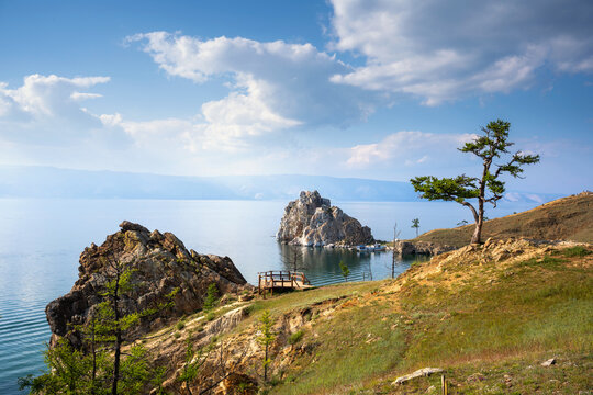 Landscape of Lake Baikal and Olkhon Island. Summer view of rocky coast, lone tree, wooden observation deck, and nature Shamanka Rock in the distance. Travel destination in Siberia, Russia