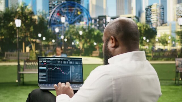 Man reads financial data graphs on a forex web platform using a laptop in a sunny green park, reviewing candlestick charts, market analysis trends and foreign exchange investing information outdoors.