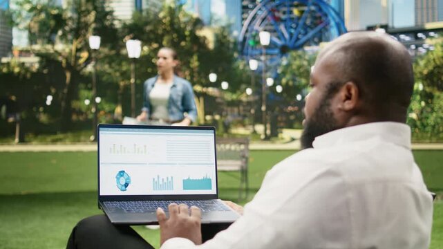 Businessman uses a laptop displaying a financial dashboard in a sunny park, reviewing data analytics, performance metrics and trading chart indicators on the screen during an outdoor work session.