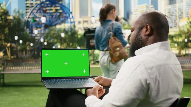 Green screen chroma key appears on a laptop held by a businessman in an urban park, providing a replaceable widescreen display for website or business content.