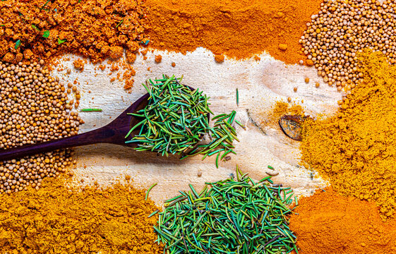 Colorful assortment of aromatic spices on a rustic wooden background, featuring rosemary in a wooden spoon, surrounded by mustard seeds, turmeric, curry, and paprika powders. Flat lay composition.