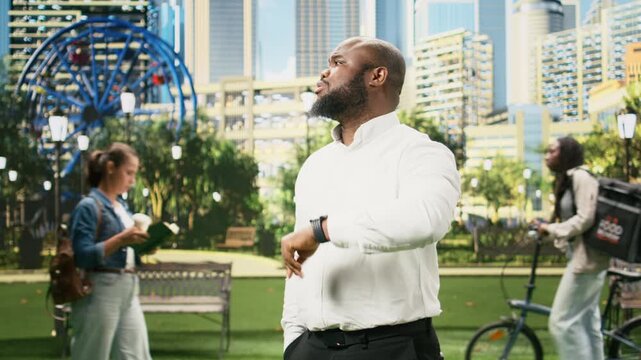Worried businessman looks down at a smartwatch in a city park, impatiently checking the time and schedule during a busy day of remote work.