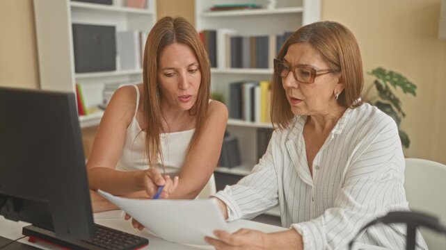 Woman points pen to document at computer while older woman holds papers and looks concerned in office building; concern collaboration.