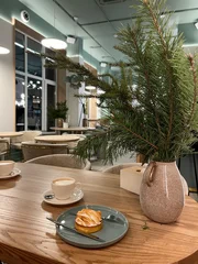 Fotobehang Horeca modern cozy cafe interior with a cup of coffee, small pastry with cream on a plate, and a decorative pine branch in a ceramic vase on a wooden table. Warm minimalist atmosphere, elegant Scandinavian  © Olena Avramenko 