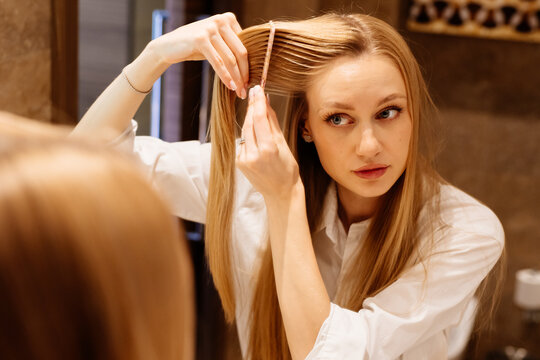 Woman preparing long hair beside lighted bathroom mirror with care. A waist-up view emphasizes grooming and preparation.