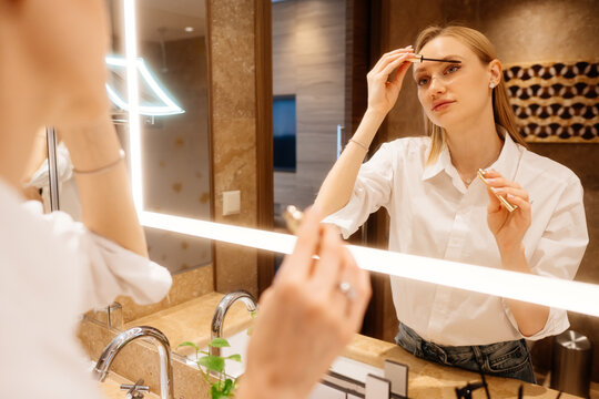Woman completing makeup with mascara before lighted mirror with care. The final grooming step completes her getting-ready process.