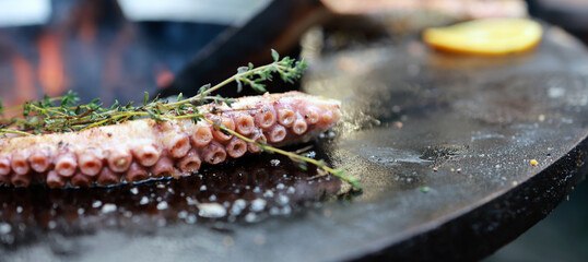 An octopus is placed on a grill over open flames. Fresh herbs and lemon slices are added for flavor. This takes place outdoors. © Anton Dios