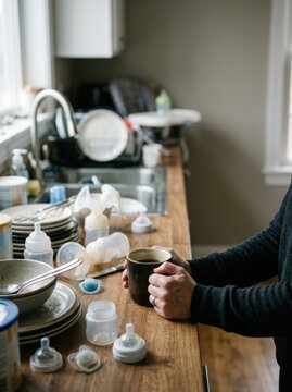 Exhausted parent holding a cup of coffee amidst a pile of dirty dishes and baby bottles in a cluttered kitchen