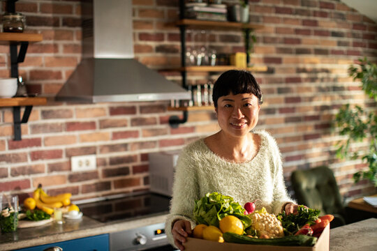 Smiling woman with box of fresh vegetables in home kitchen